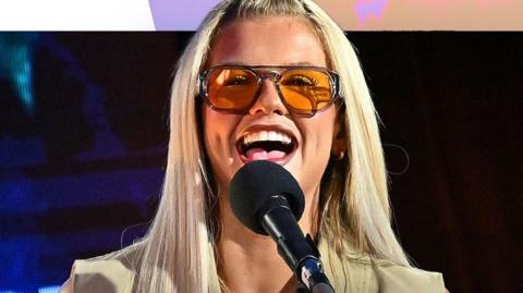 Skye Newman smiles behind a mic. She has long blonde hair and wears orange sunglasses