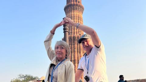 A woman in a beige jacket holds her right arm above her head. A man in a white t-shirt holds his left arm in the air and holds her right arm. They are in front of a tower.