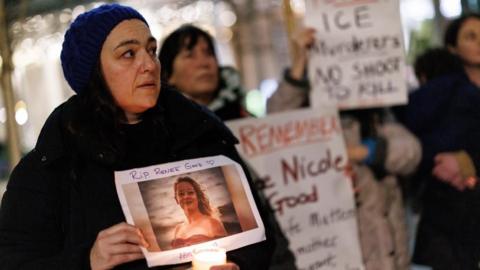A woman wearing a dark jacket with a hood over her head holds a candle and a picture of a Minneapolis woman fatally shot by an agent of ICE, the US Immigration and Customs Enforcement agency