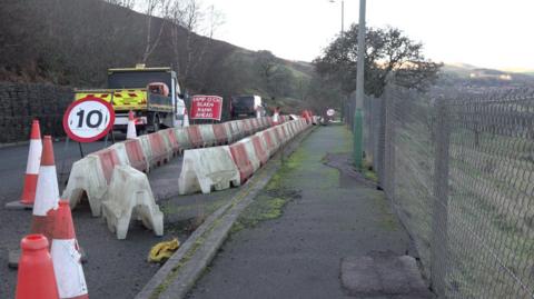 The A469 with visible damage to the road and pavement. Red and white traffic bollards and cones stretch along one side of the carriageway. Some vehicles are traveling along the one side of the road. In the background are trees along the hillside and there's a metal fence on the right side of the picture.