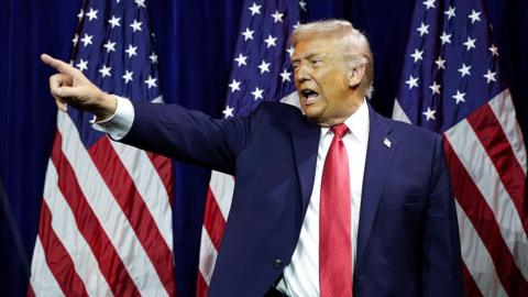 US President Donald Trump, wearing a blue suit and red tie, points with his finger in front of an American flag.