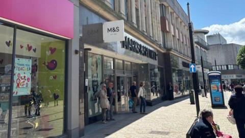 A row of stores going from left to right on the photo, including Marks & Spencer, with those words being above the door. Some people are standing in front of the store.