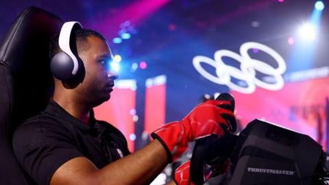 A man wearing red gloves has his hands on the wheels of a driving simulator and wears headphones. In the background are the olympic rings in lights.