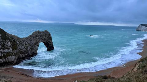 The arch of Durdle Door seen from a high vantage point with the beach in the foreground and a choppy-looking sea in the background beneath cloudy skies.