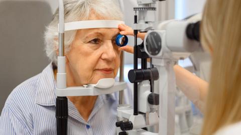 An older lady having an eye test. She is looking into a machine while an optician hold a lens to her left eye.