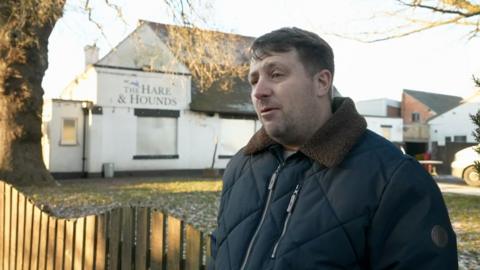 A man with brown hair wearing a blue padded coat with brown collars outside a closed pub. The building is white with The Hare & Hounds written on the wall
