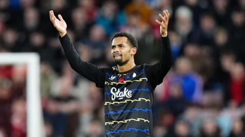 Iliman Ndiaye wears a black jersey with blue and yellow zig-zag stripes. His arms are raised in celebration after scoring a goal.