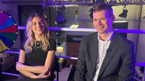 A female reporter and a male reporter look at the camera smiling