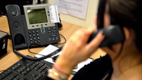 A woman uses a landline phone as she sits at a desk. There is a keyboard in front of her. The view is from behind her shoulder so her face is not visible but she has dark hair tied back and has bangles on her wrist.