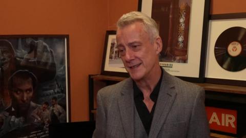 Stephen Tompkinson looking to the left of the picture and slightly downwards. He has grey hair which is swept back slightly. He is wearing a grey tweed blazer and a black shirt. He is smiling as he talks. He is standing in front of an orange wall with various posters.