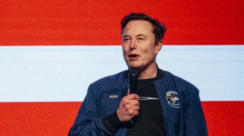 A man in a blue jacket that reads "Musk" speaks into a microphone, standing in front of a red and white striped backdrop.