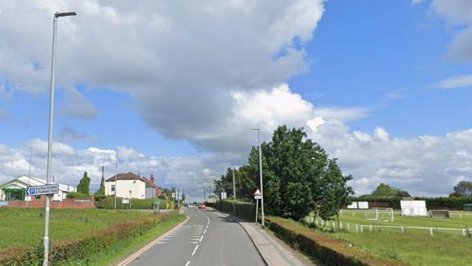 A rural road with fields on both sides. Lamp-posts and trees can also be seen.