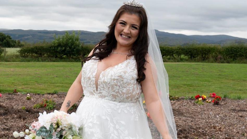 Melissa Ashcroft wearing a white lace wedding dress holding a bouquet of flowers and wearing a silver tiara and veil. She has brown curled hair and is smiling at the camera. Green fields and rolling hills are in the background.