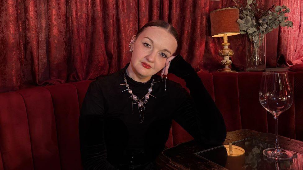 A woman with a black jumper and decorative necklace sits at a table at a bar with a large empty wine glass. The sofa and curtains behind it are red