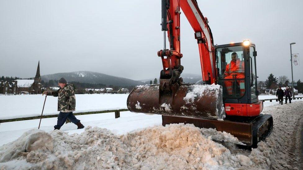 An orange digger clearing deep snow in Aboyne in Aberdeenshire as people walk past.