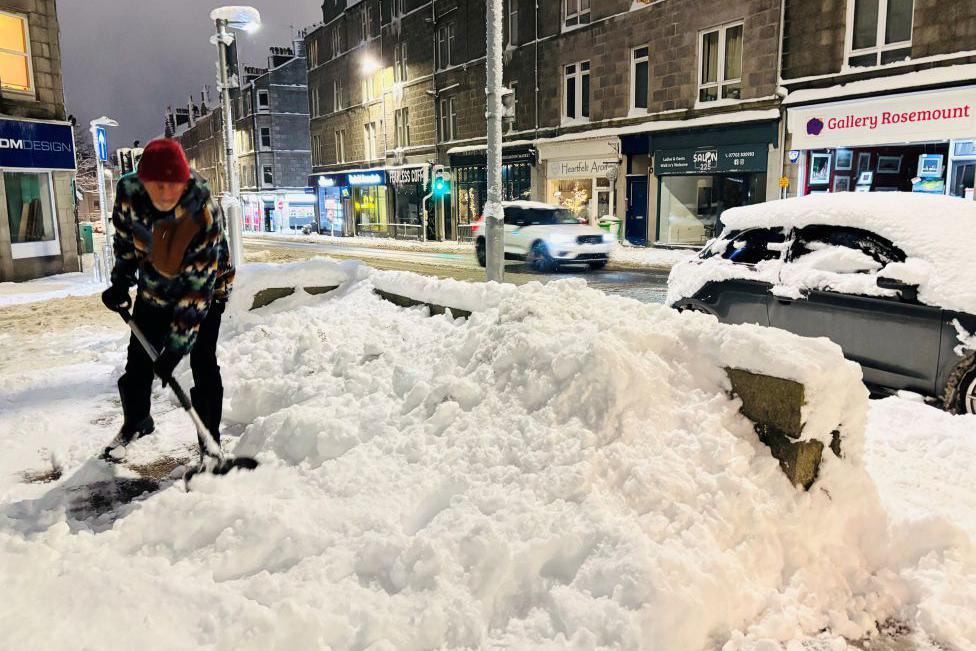 A man using a shovel to clear snow in Aberdeen, with snow-covered cars in the background.