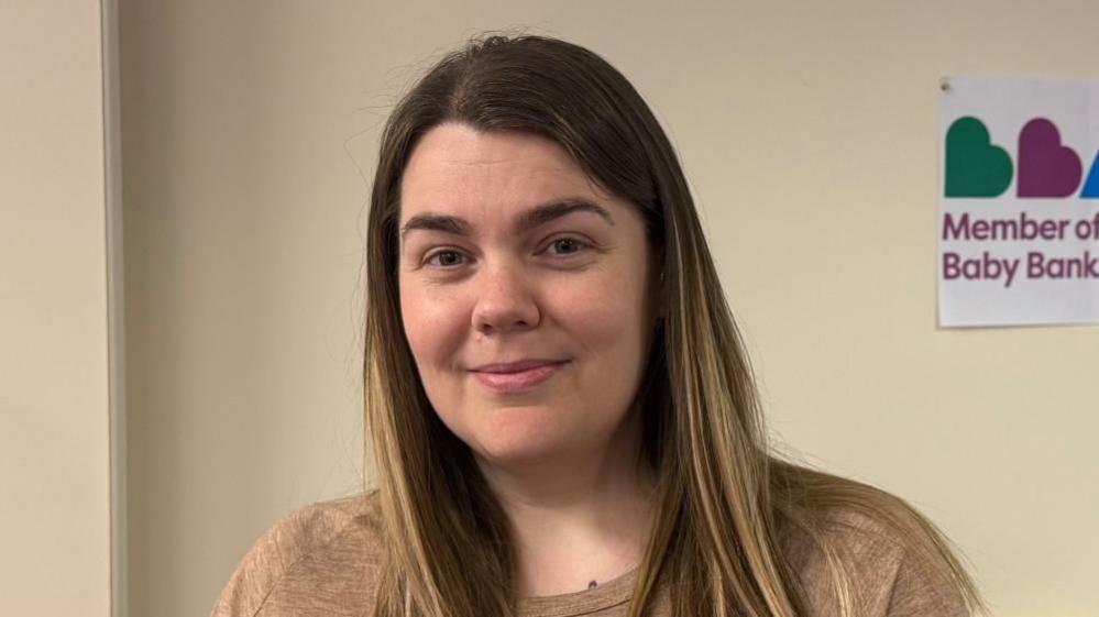 Elise McKenna is looking into the camera and smiling. She has long dark brown hair and is wearing a light brown top