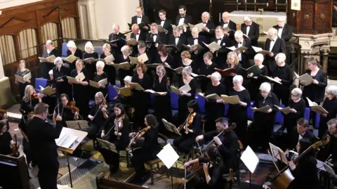 A previous concert by Wombourne and Distrrict Choral Society. There are four rows of singers, who are all wearing black clothes. In front of them are musicians playing sting instruments and a conductor. 