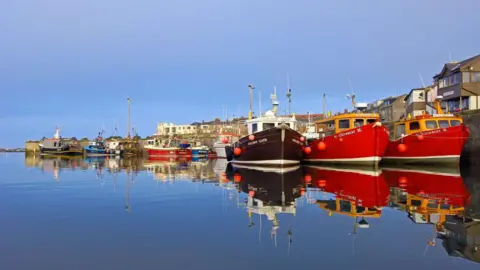 Fishing boats moored at the harbour at Seahouses. Their reflection can be seen in the water. There are three berthed in the foreground with more behind. Set back from the harbour wall on the right is a long row of houses and other buildings which face out to sea.