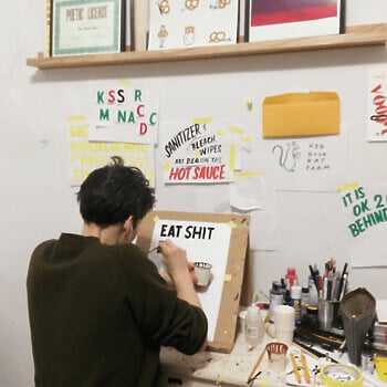 A photograph of a person painting the words “EAT SHIT” onto a piece of paper taped to cardboard that’s leaning on a desk, with other hand-lettered paintings hanging on the wall behind it.