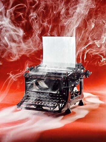 A photograph of an old-fashioned black typewriter sits on a red backdrop with smoke all around it. There’s a blank piece of paper rolled into the typewriter.